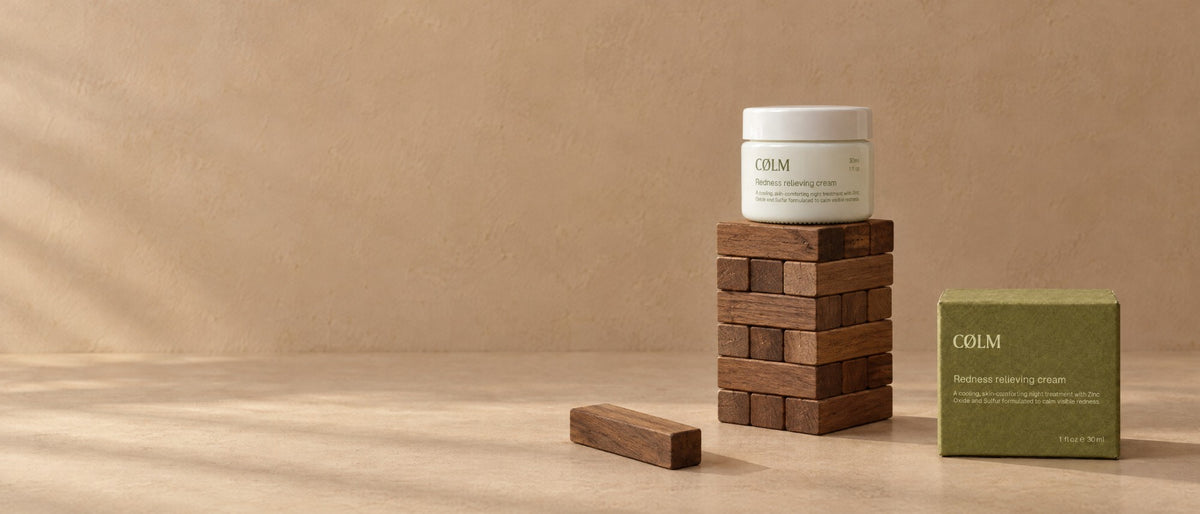 CØLM Redness Relieving Cream på toppen af et lille tårn af mørke træklodser, med den olivengrønne æske ved siden af