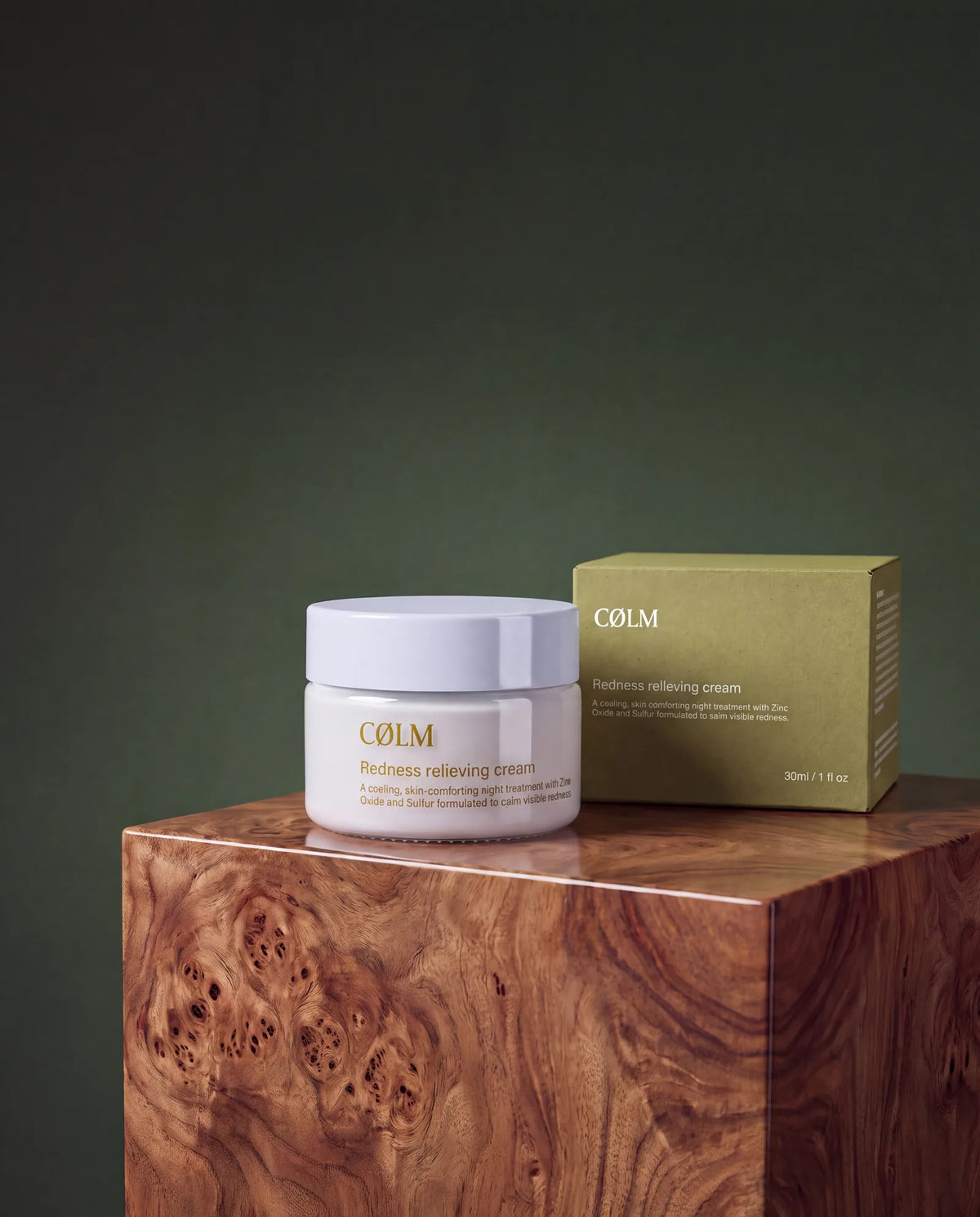 CØLM Redness Relieving Cream, bøtte og genbrugspap-æske centreret på træpodie mod mørkegrøn baggrund